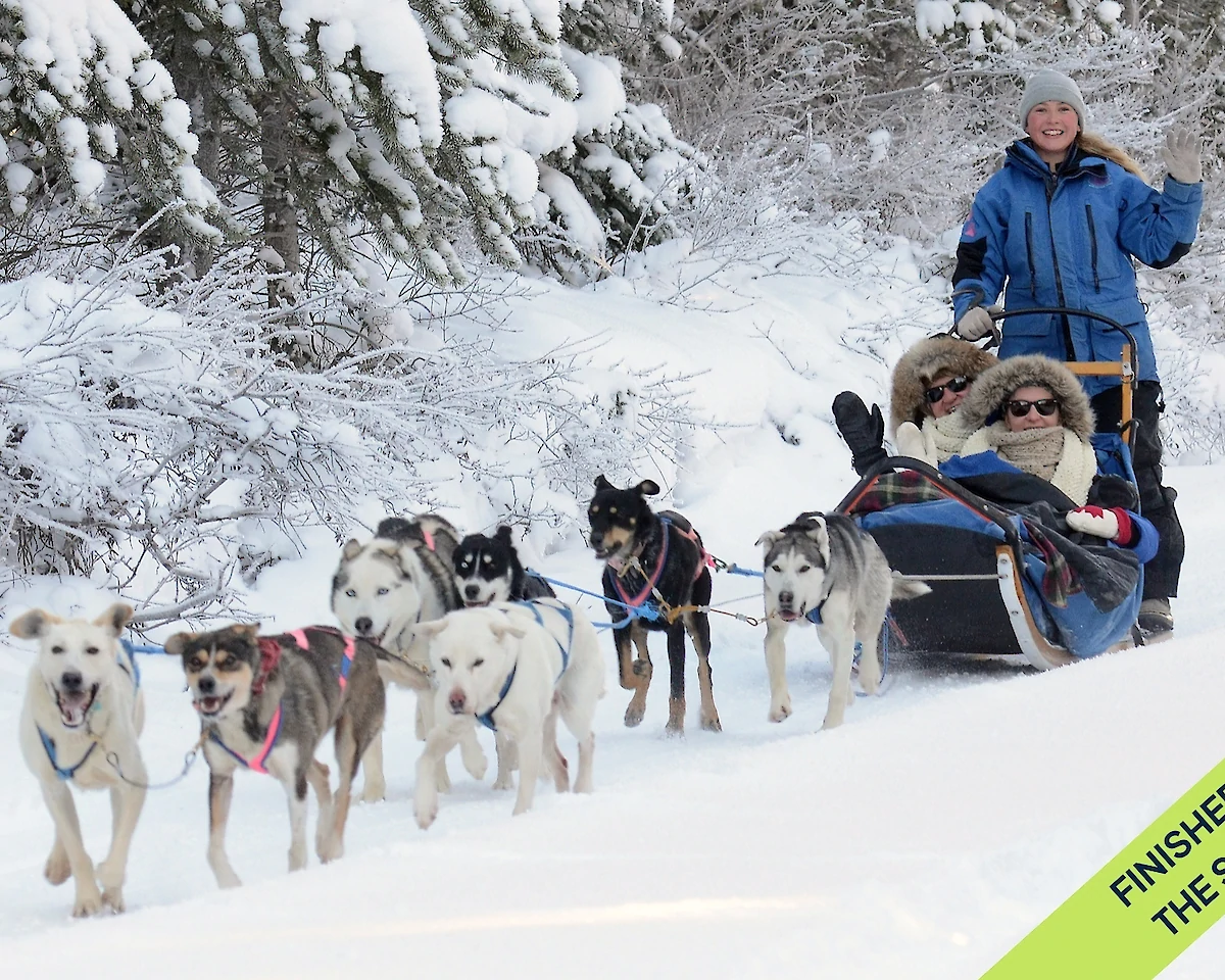 Two guests riding in a dog sled with the guide mushing behind.