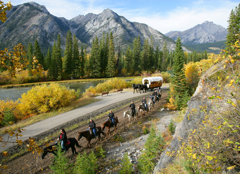 People riding horses and horses pulling a wagon along the Bow River in Banff to the Cowboy cookout