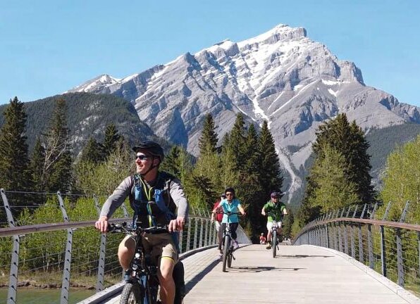 People cycling Ebikes over the pedestrian bridge in Banff with Cascade Mountain in the background